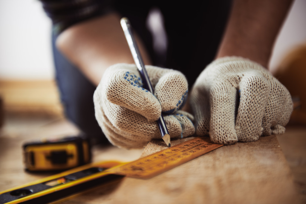 close up of craftsman hands in protective gloves measuring wooden plank with ruler and pencil